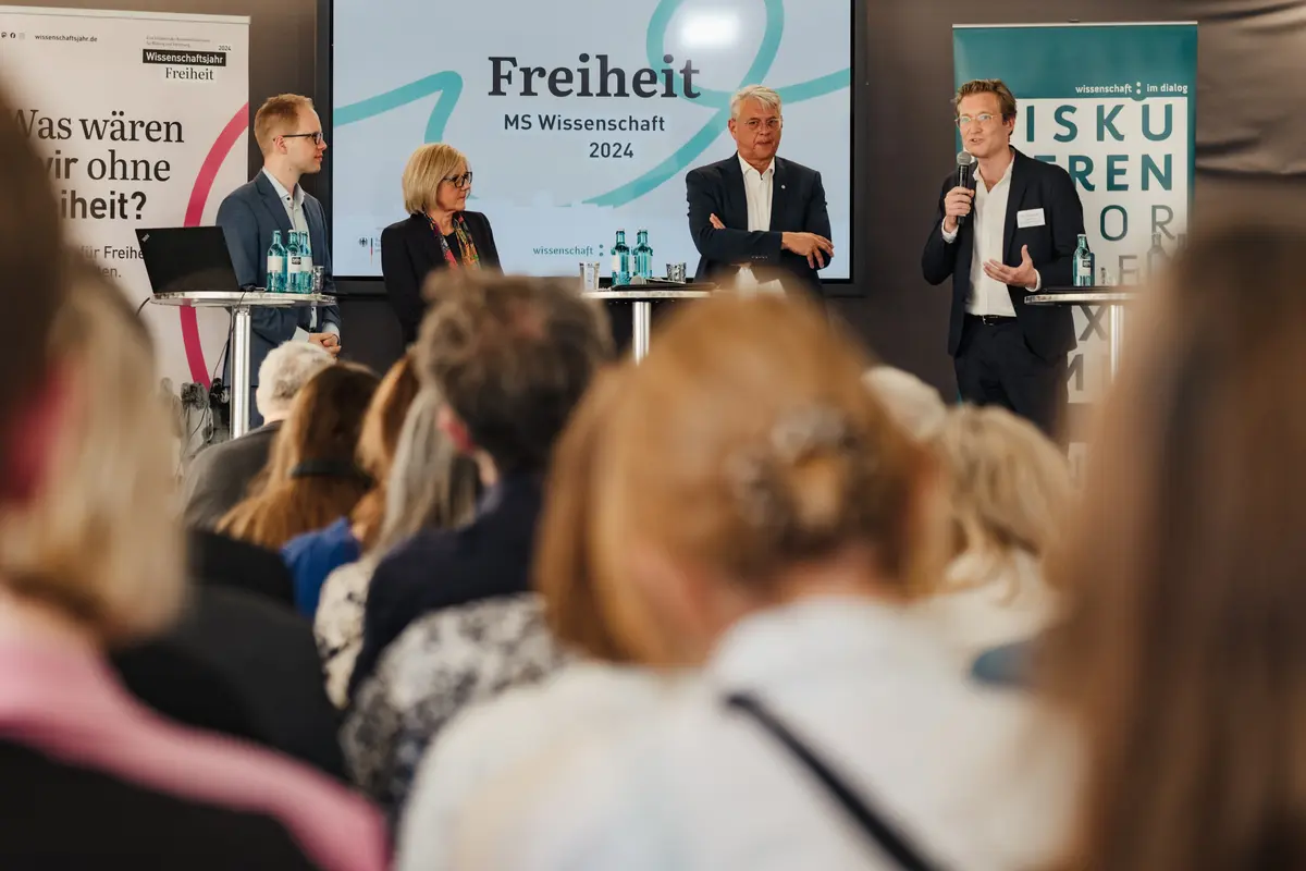 Eine Pressekonferenz. Im Vordergrund sieht man Zuhörende sitzen, weiter hinten im Bild sieht man das Podium: Drei Männer und eine Frau.